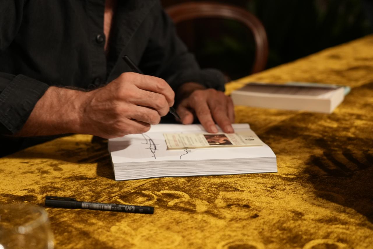 A hand signing the title page of the book, a marker on the table
